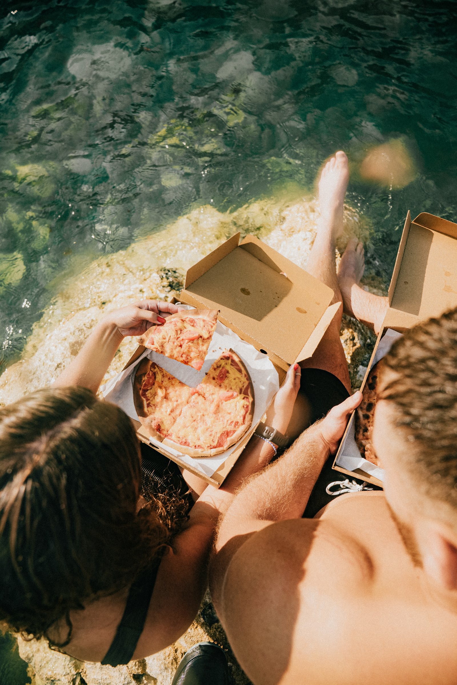 A couple sitting on a rock by the water, eating pizza