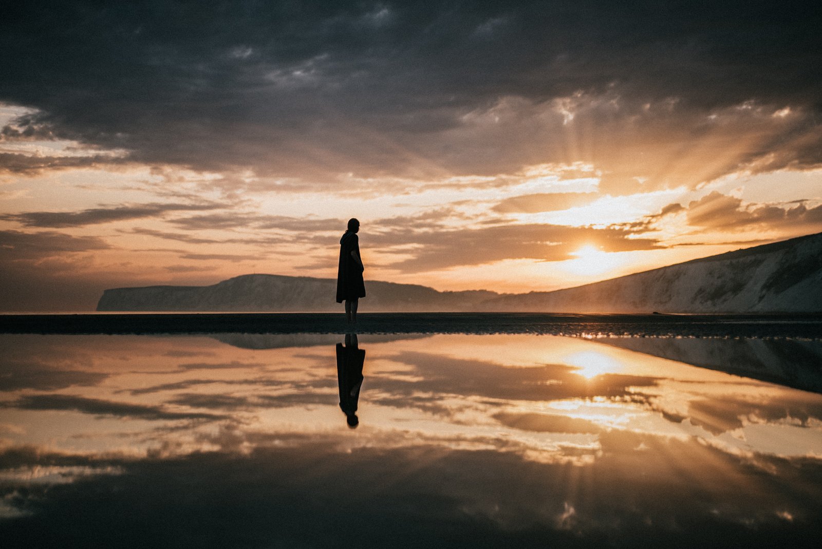 A woman stands on a beach at sunset, looking out to sea